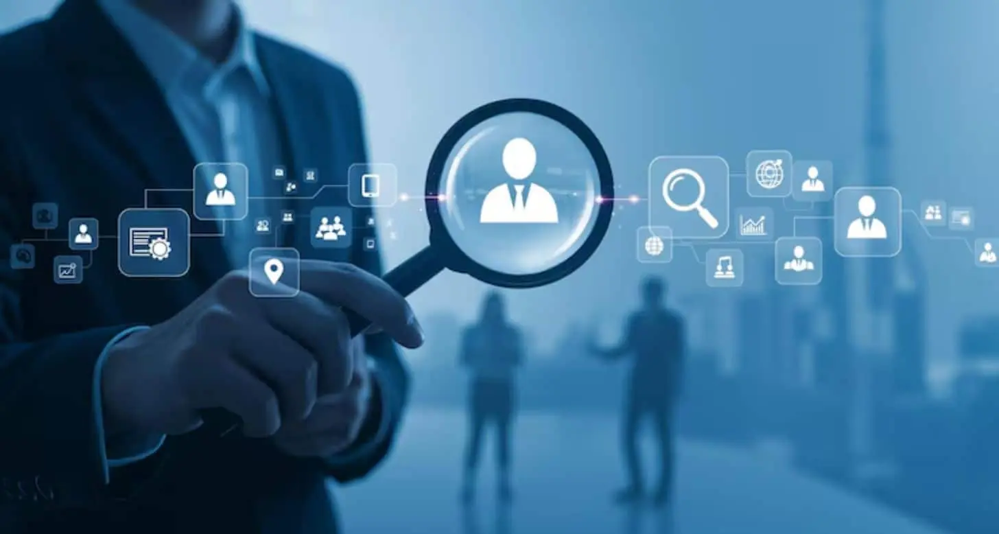 Business professional holding a magnifying glass highlighting a candidate profile, with digital recruitment icons representing talent search, hiring, and workforce management.